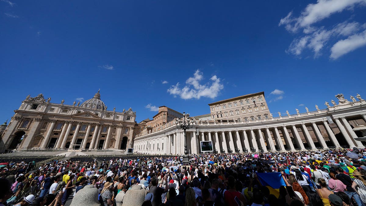 A large crowd gathers at St. Peter's Square in front of the Vatican with clear skies overhead