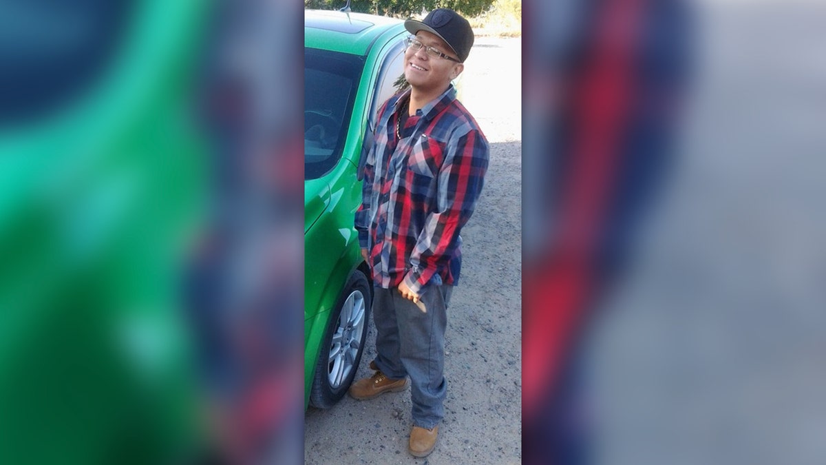 Zachariah Shorty wearing a blaid shirt, jeans and workboots standing next to a green car