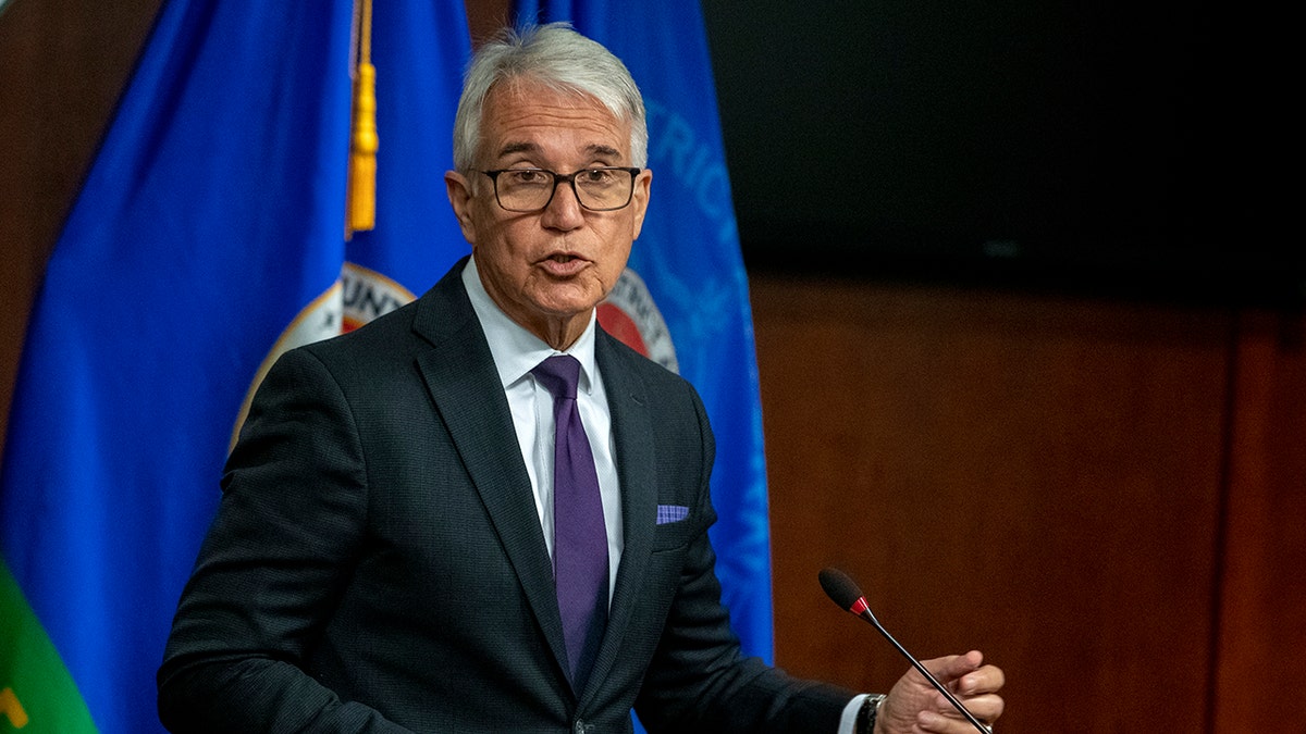 Gascon speaking in front of a microphone, wearing a suit and tie and a flag behind him