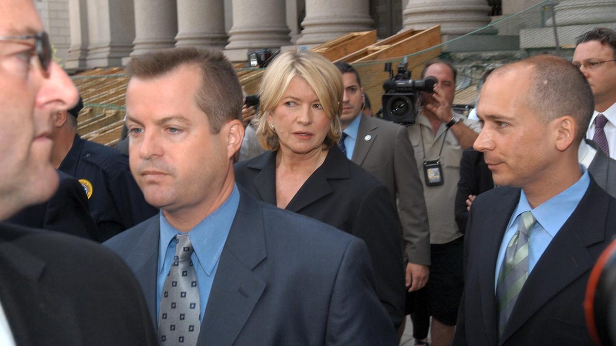 Martha Stewart in a black suit looking perplexed in a crowd of lawyers and reporters after being sentenced to prison