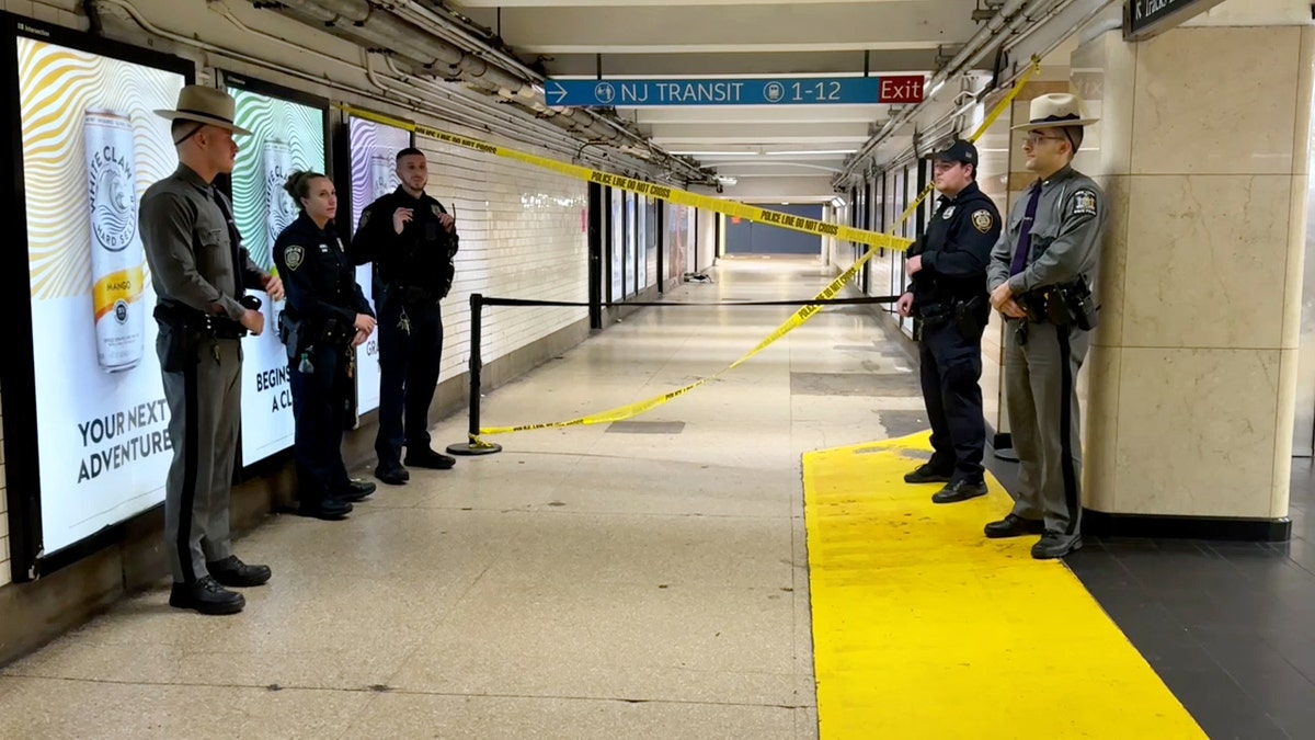 Five New York police officers stand guard outside of a Penn Station hallway cordoned off with caution tape.