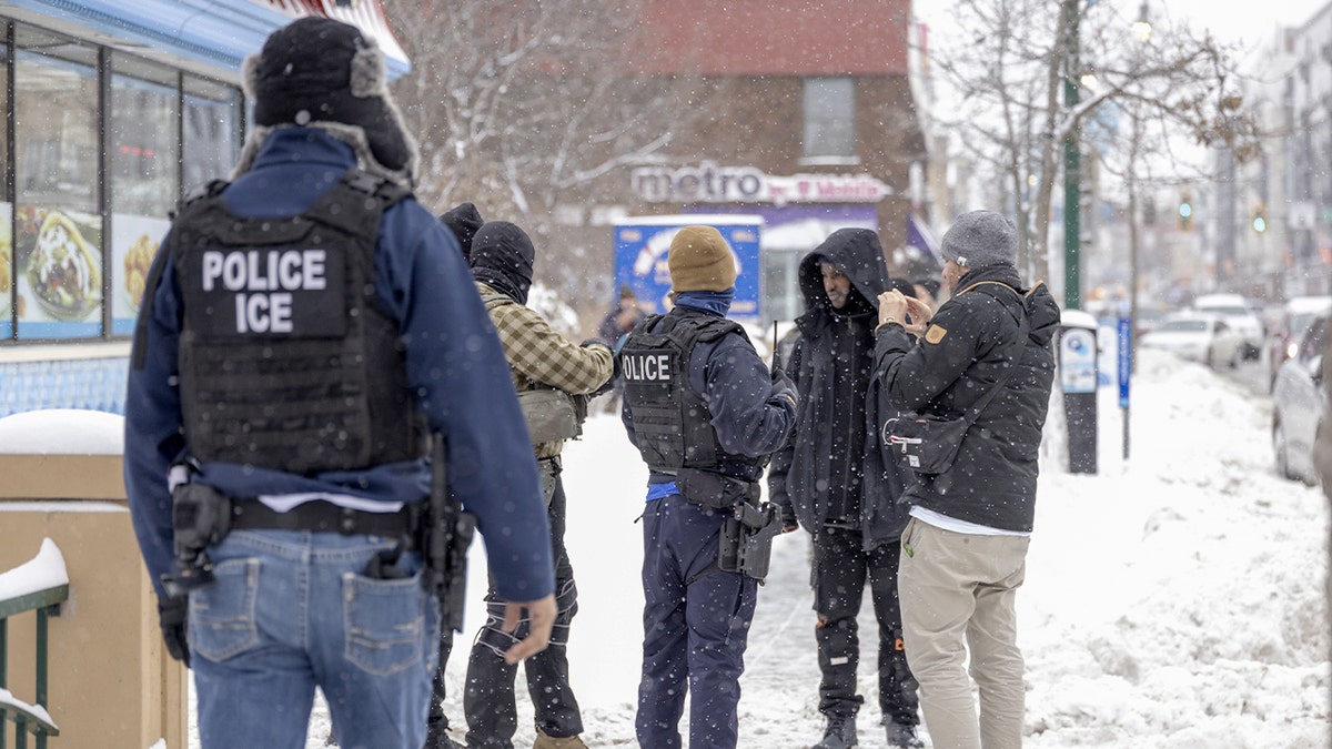 Federal officers speak with a man on a busy commercial street as bystanders look on.