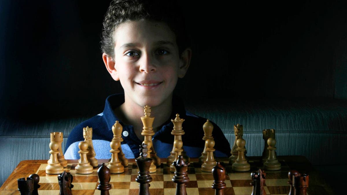 A young Daniel Naroditsky smiles behind a chessboard; the chess grandmaster has died at 29.