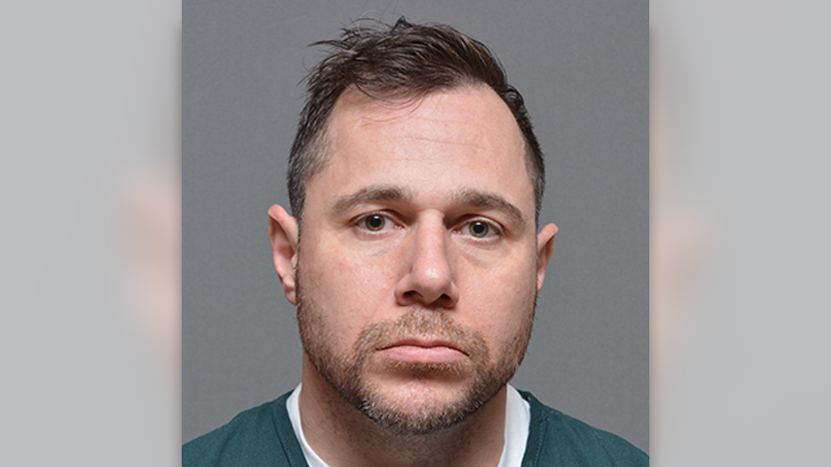 Paliccio has a stubbled beard and unkempt hair as he poses for a mugshot in a green jail jumpsuit over a white T-shirt