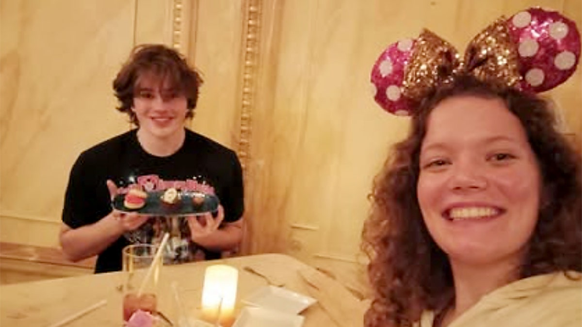 A girl takes a selfie while wearing Mickey Mouse ears, and a boy next to her holds a cake.