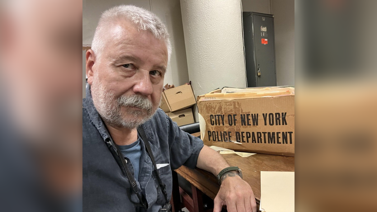 Vronsky in a shirt at a desk with a box that says "City of New York Police Department"