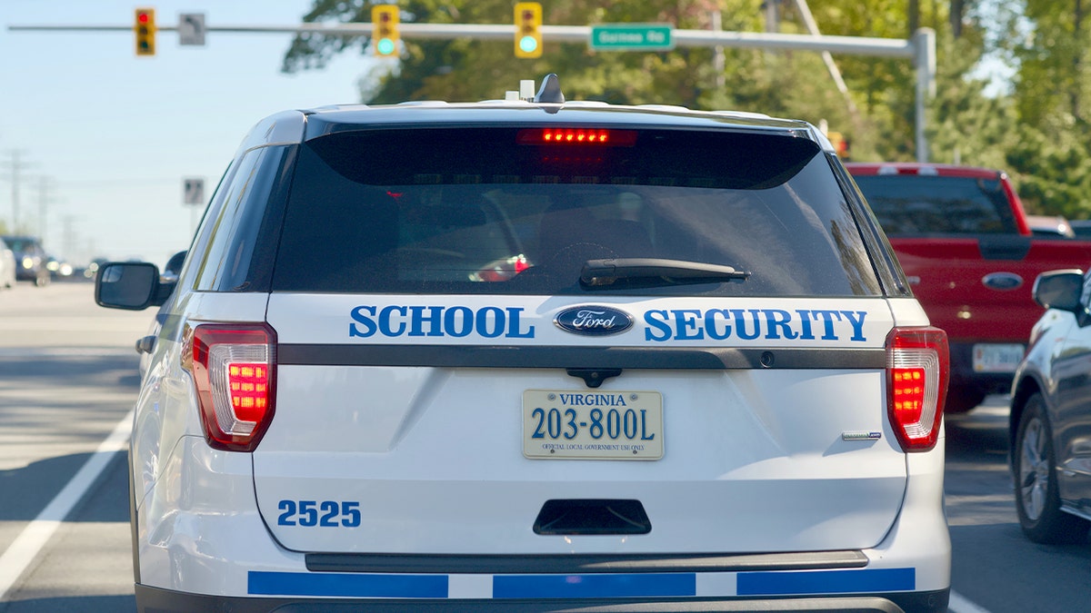 A marked school security vehicle pauses at a suburban intersection near a school campus.