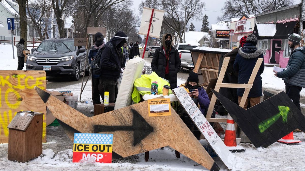 Masked people stand by a wooden barricade with signs while vehicles wait on a snowy street.