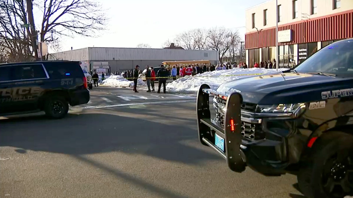 People standing behind a taped off portion of the road with police cars and a school bus in view