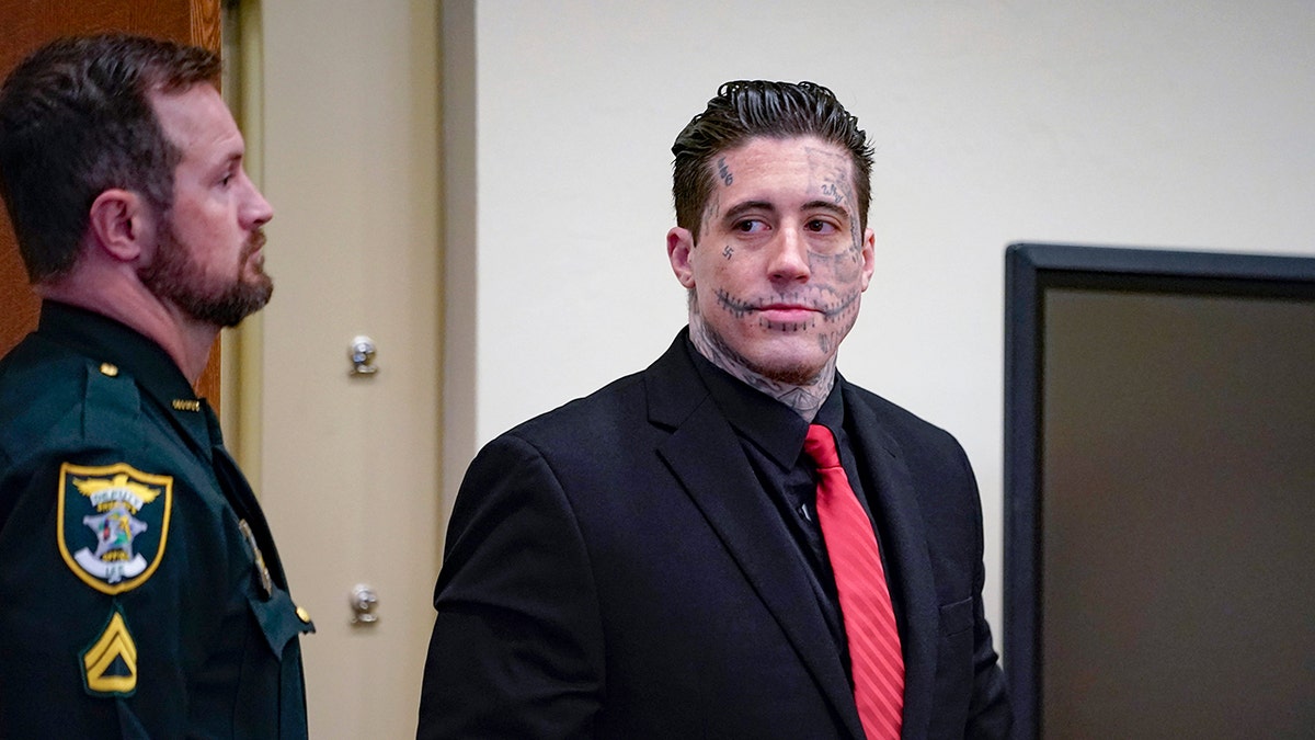 Wade Wilson wearing a dark suit with a red tie next to police.