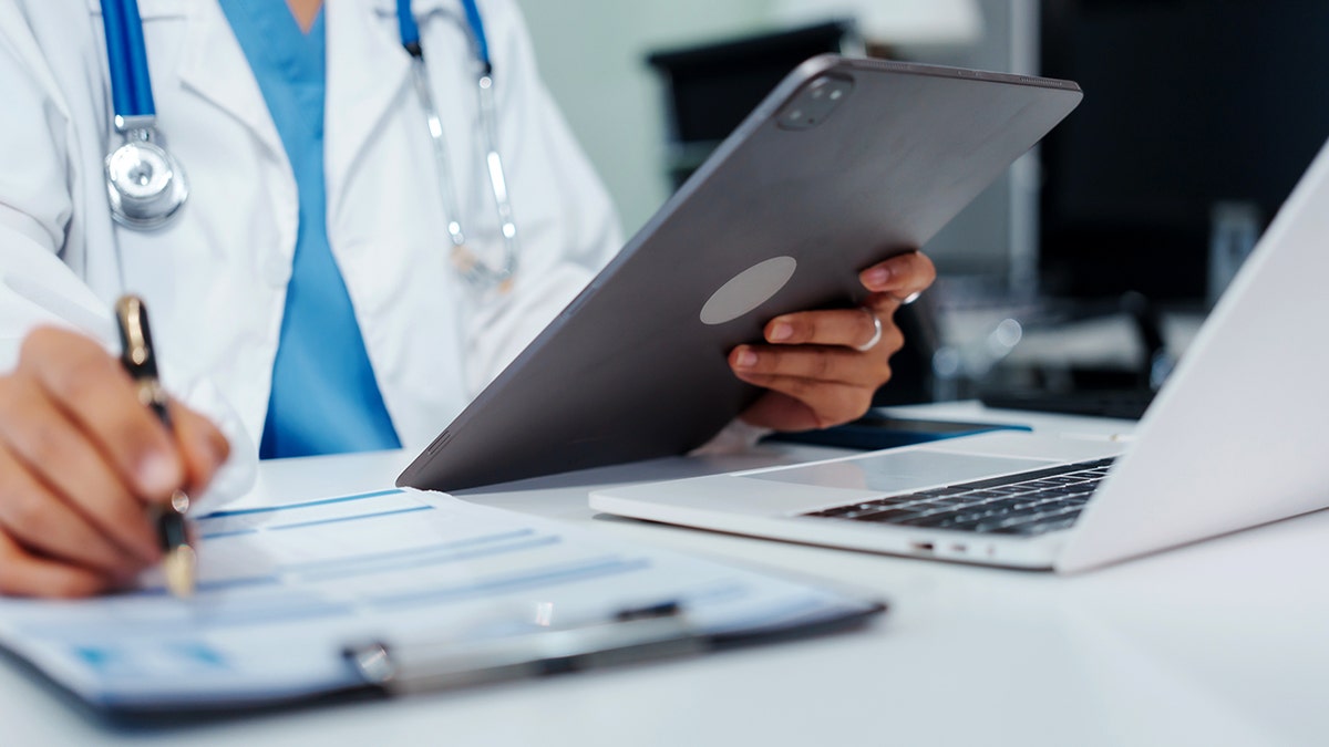 A doctor looking at a tablet and writing on a clipboard.