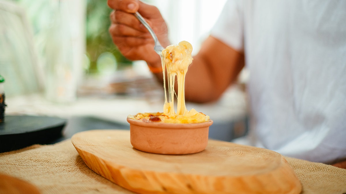 Young man lifting a forkful of cheesy macaroni and cheese from a small terracotta bowl.