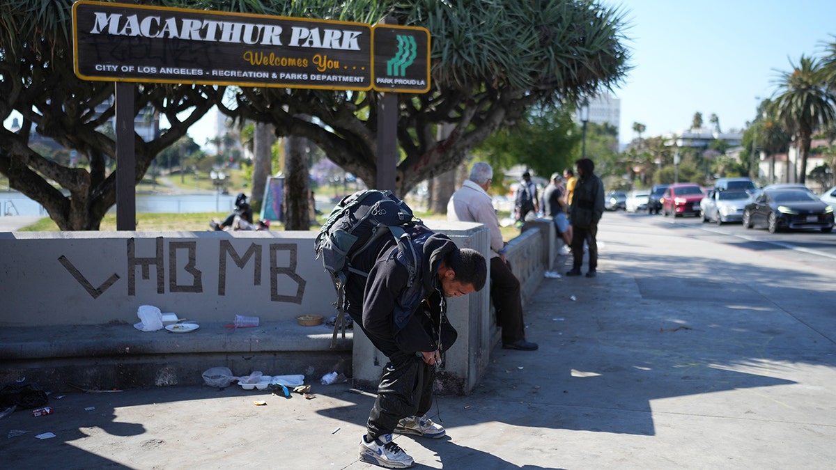 A man holding a pipe hunched over near a MacArthur Park welcome sign in Los Angeles