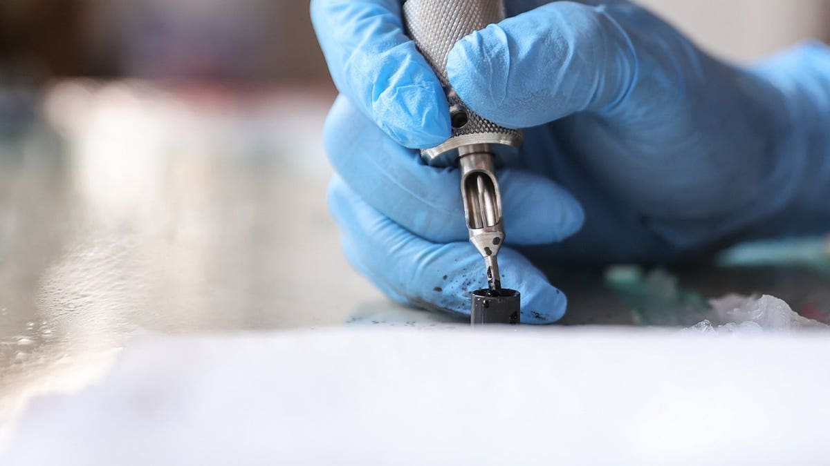 A hand wearing a blue glove holds a tattoo machine