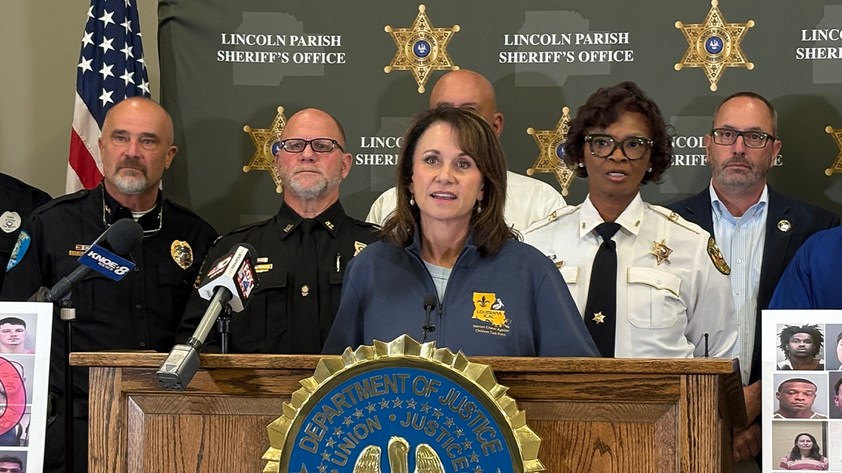 Liz Murrill in a blue windbreaker speaking behind an oak podium at a press conference