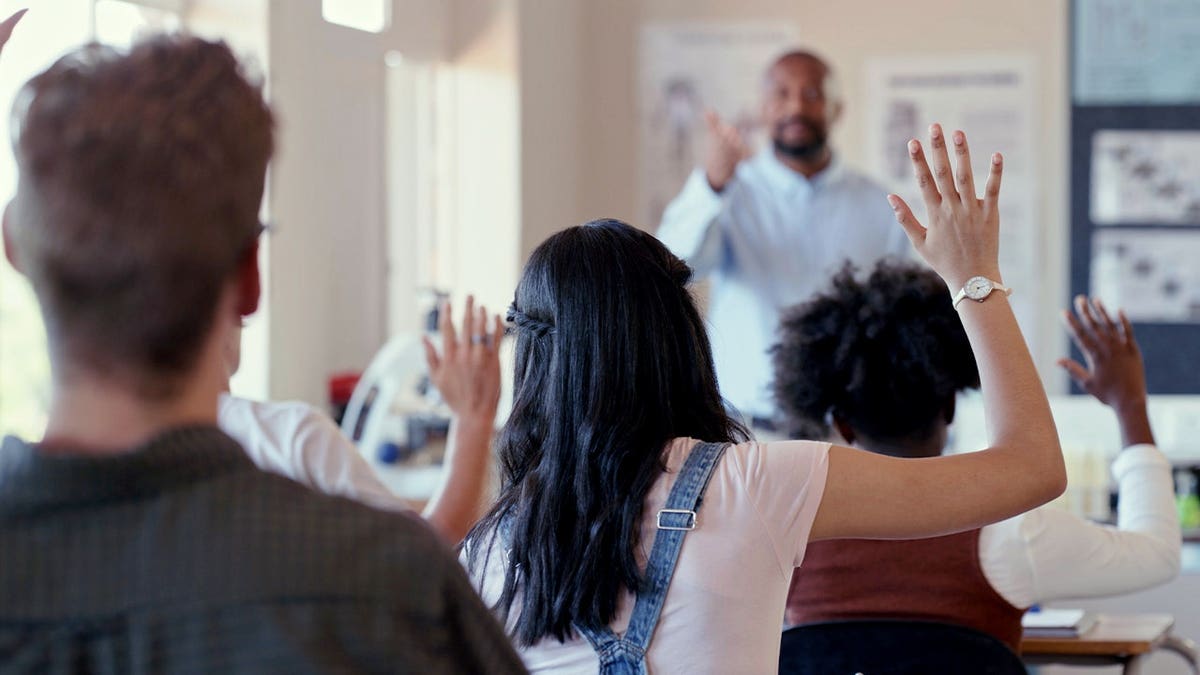 File photo showing students raising their hands in a classroom while a teacher stands at the front of the room.