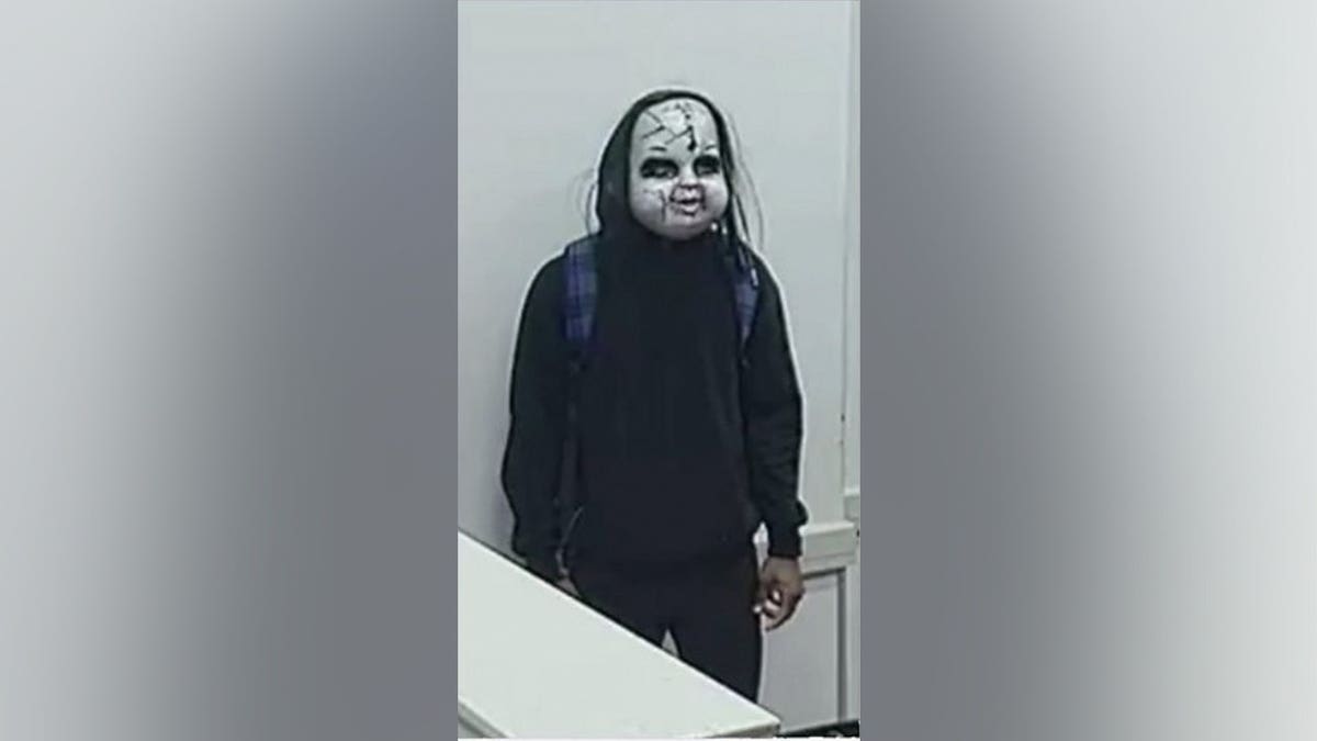 Surveillance still of a masked person in dark clothing with a blue plaid-strapped backpack standing against a white wall.