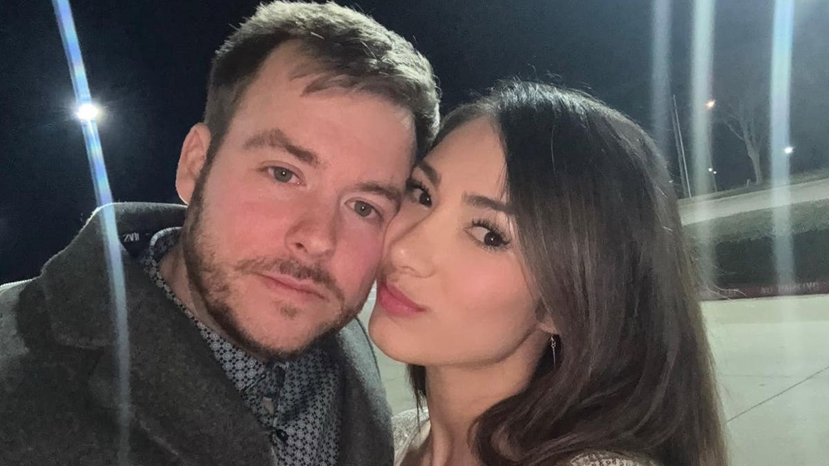 A man and a woman pose cheek to cheek for a nighttime selfie in a parking lot.