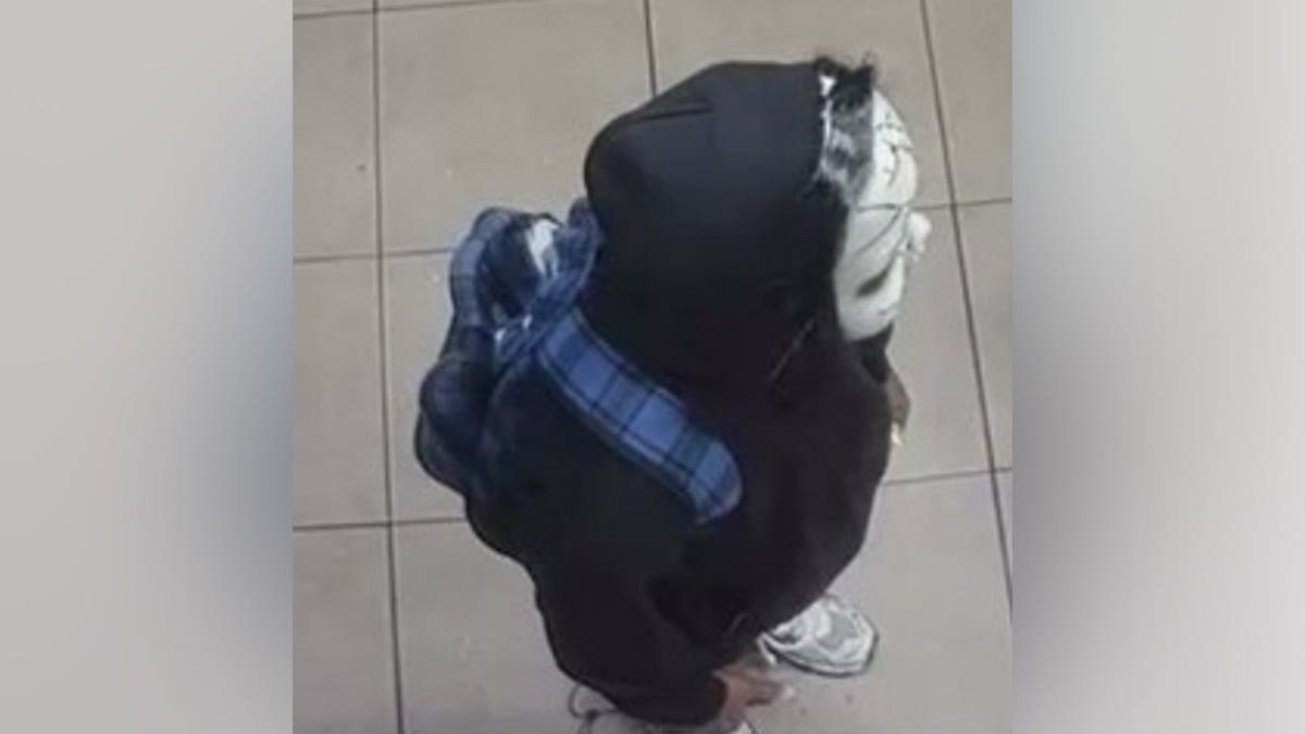 Overhead surveillance view of a hooded person wearing a doll mask and a blue plaid-strapped backpack on a tiled floor.
