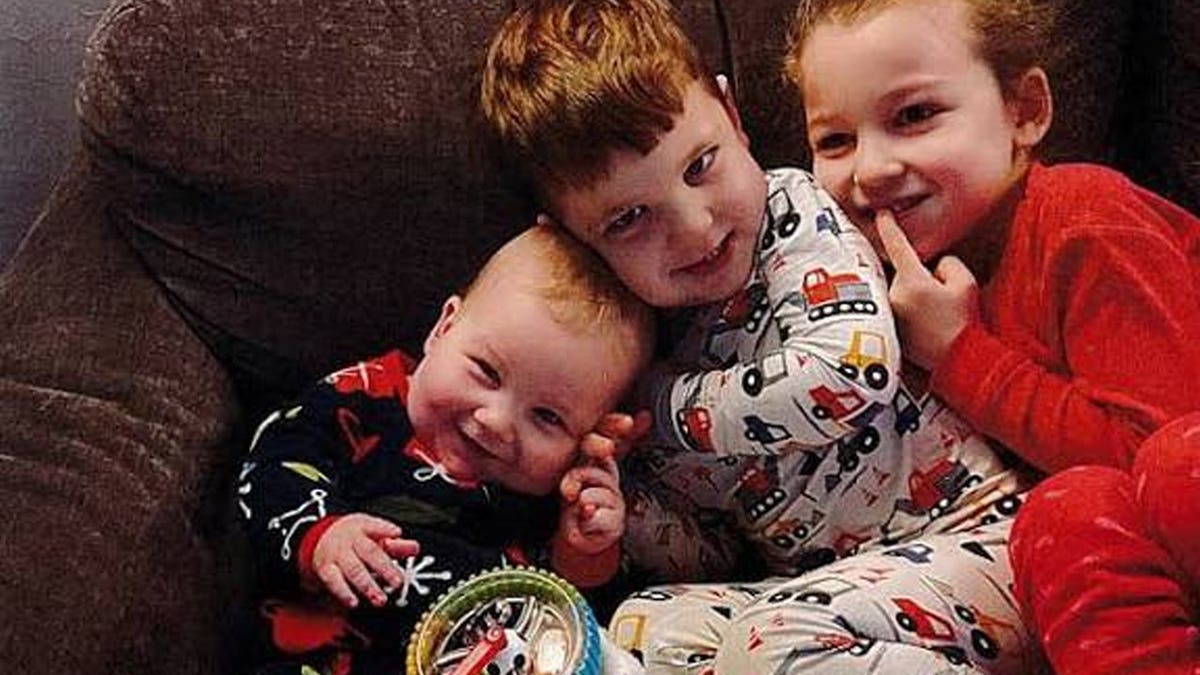 Three young Clancy children in pajamas sit together on a couch, the smiling baby resting against his older brother, with their sister beside them.