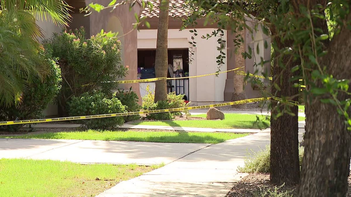 Yellow crime scene tape strung across the front walkway and entrance of a Southwestern-style home surrounded by trees and shrubs.