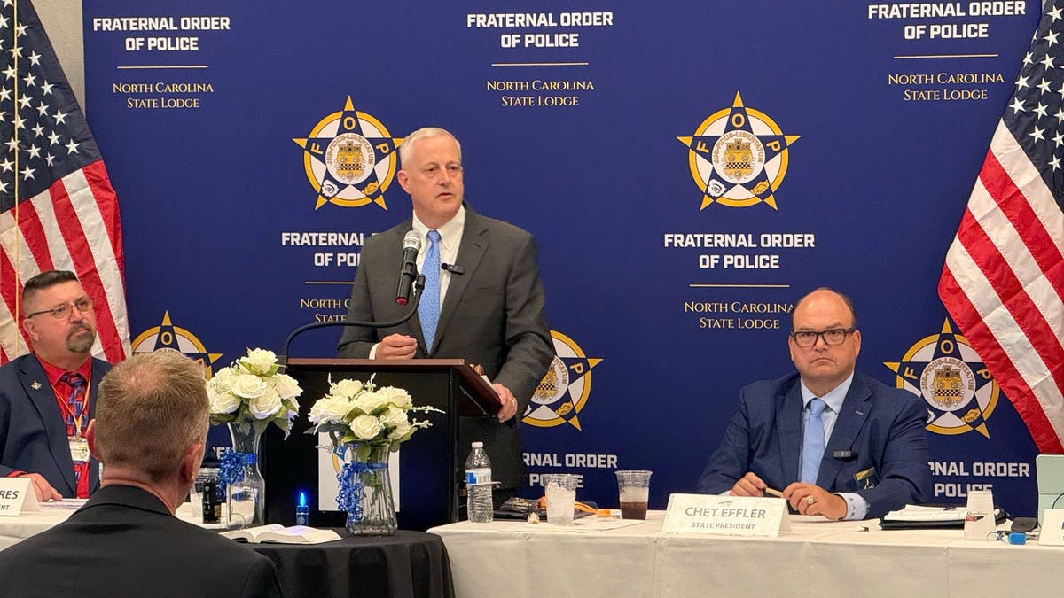 Michael Whatley speaks at a podium before a North Carolina Fraternal Order of Police backdrop as Chet Effler sits beside him.