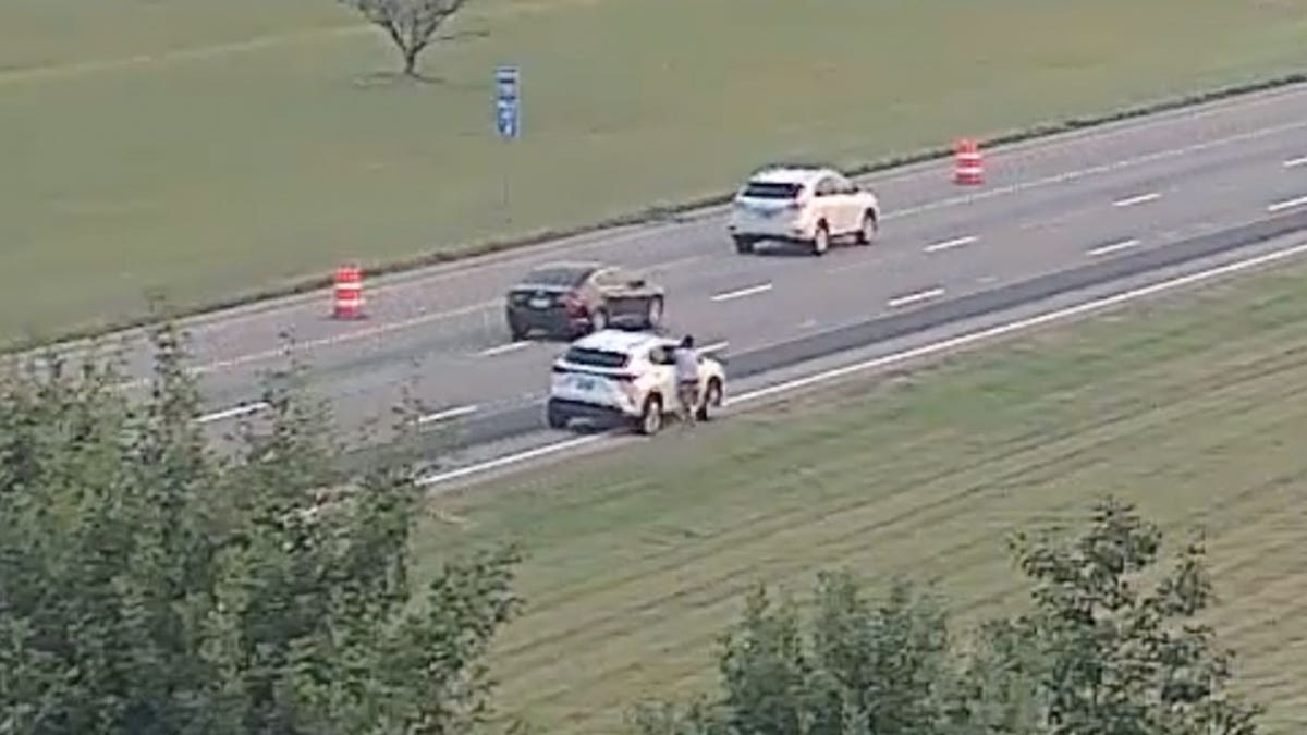 Highway camera view of a person beside a white SUV on the shoulder of an interstate near orange construction barrels.