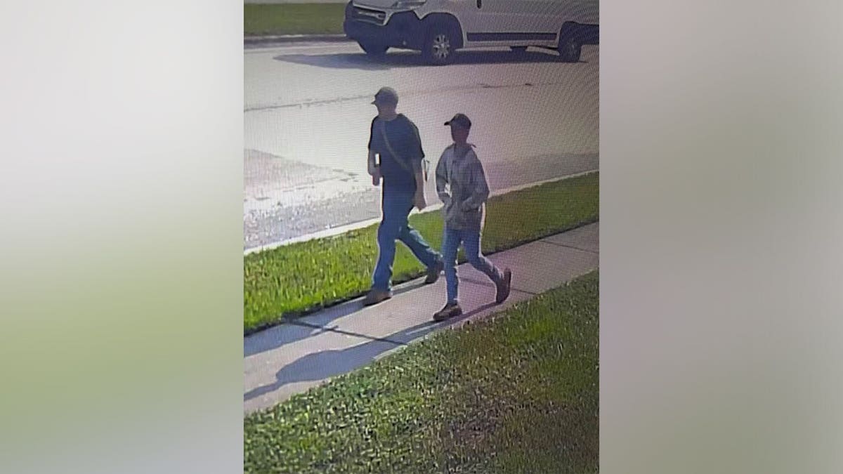 A grainy surveillance still of two young men walking along a sidewalk past a parked vehicle.