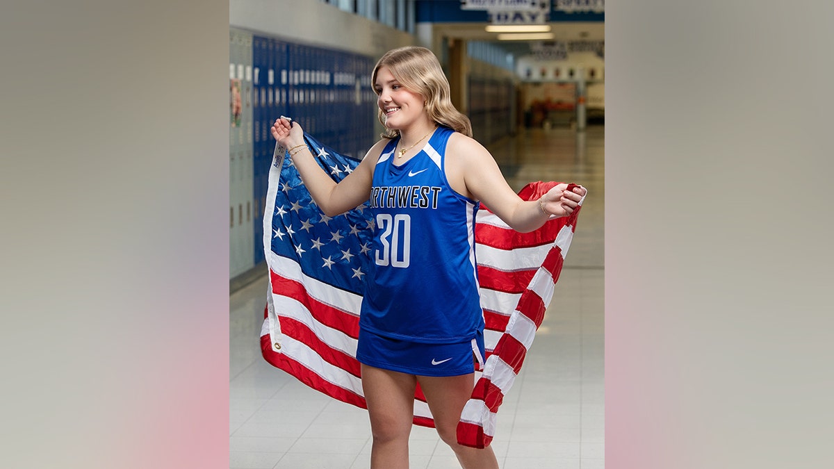 A teenage girl smiles over her shoulder, draped in an American flag over a blue athletic jersey in a school hallway.