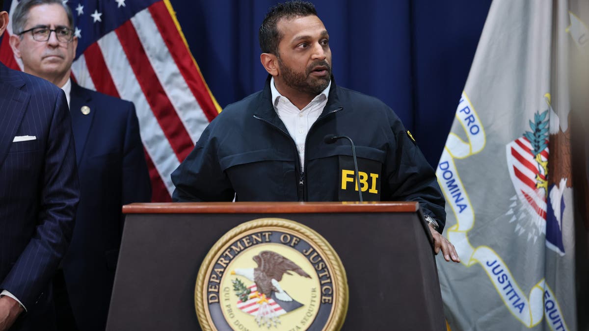 kash patel wears an black FBI jacket while standing at a DOJ podium