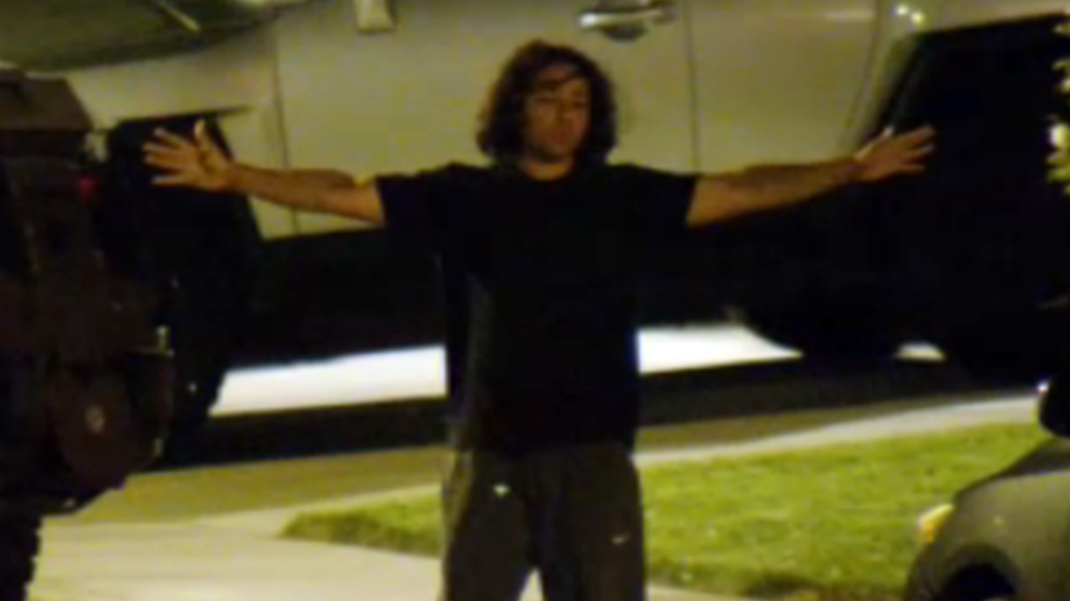 A man in a blank t-shirt with long curly hair lifts his arms as he surrenders to deputies