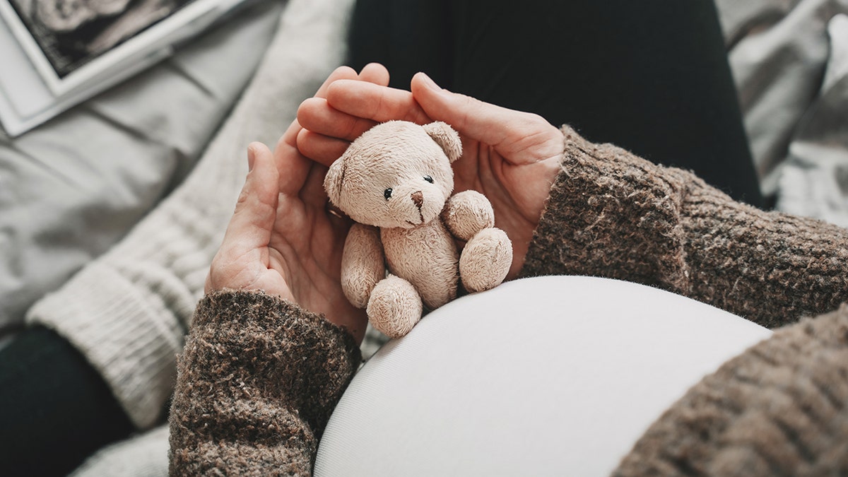 A pregnant woman holds a small stuffed toy teddy bear to her belly as she sits with her legs crossed on her bed. She cups the little toy in both hands as, out of focus on the upper left corner, there appears to be an ultrasound photo
