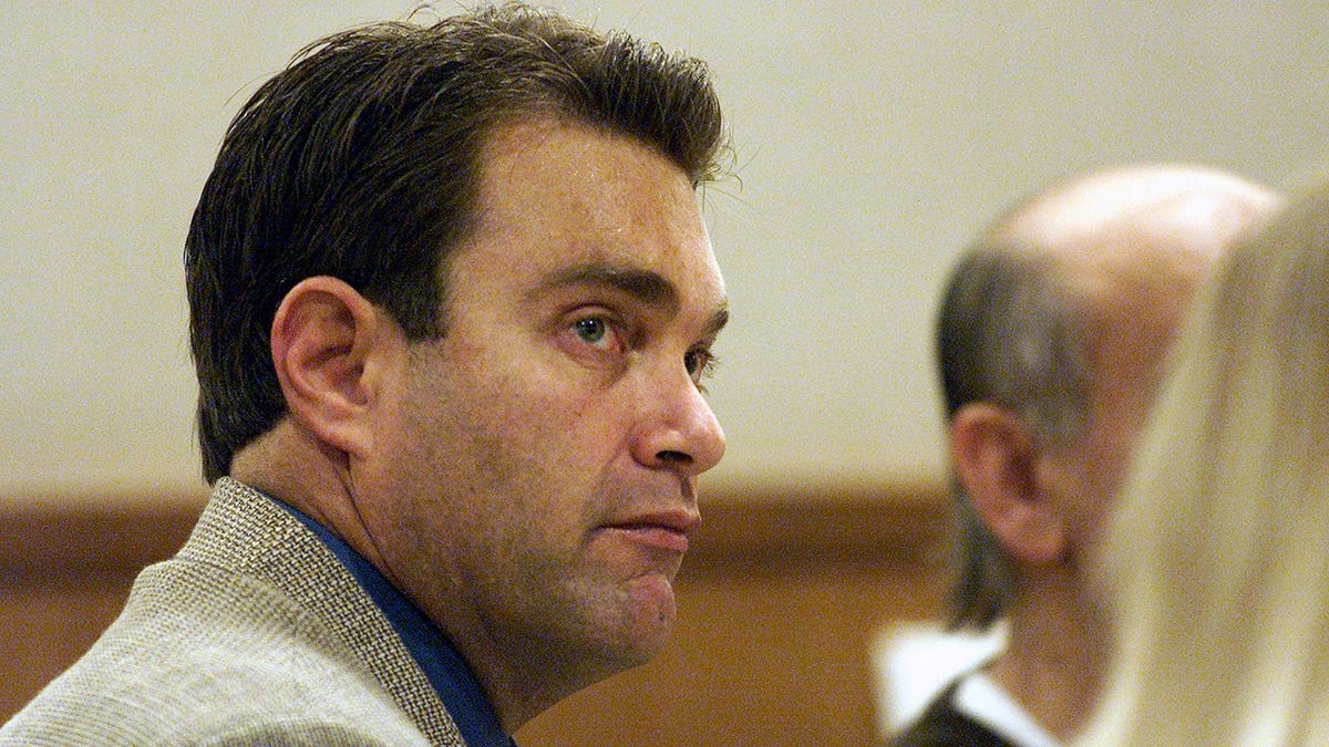 Andrew Luster, wearing a light-colored jacket over a blue shirt, looks to his right while seated in a courtroom.