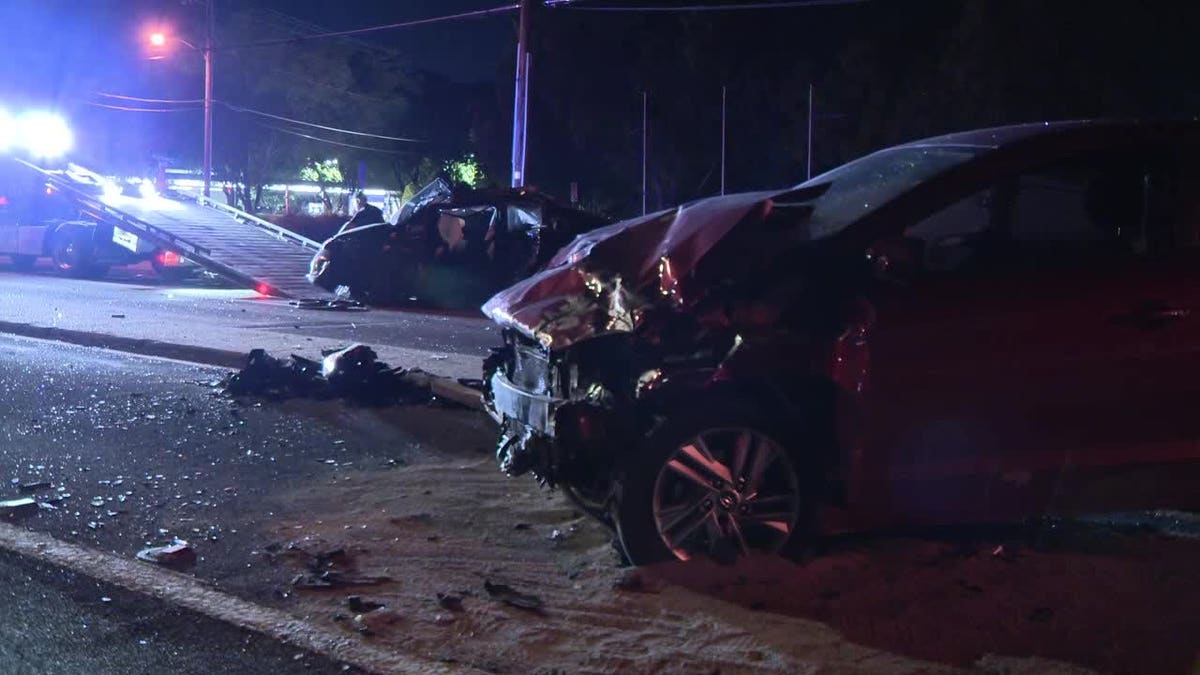 A damaged red Hyundai Elantra sits in the foreground while a heavily damaged Toyota Prius is visible near a tow truck at a nighttime crash scene.