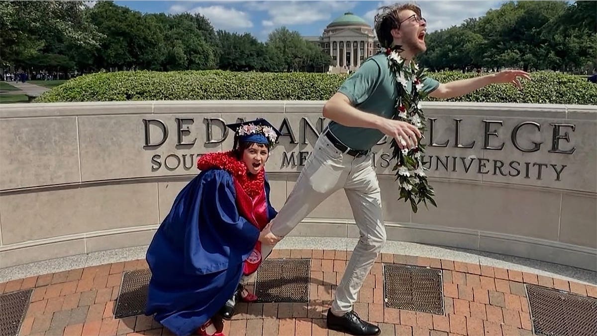 Madison Duren, wearing a blue graduation gown and cap, playfully grabs Julian Harris' leg as he poses in front of a Southern Methodist University sign.