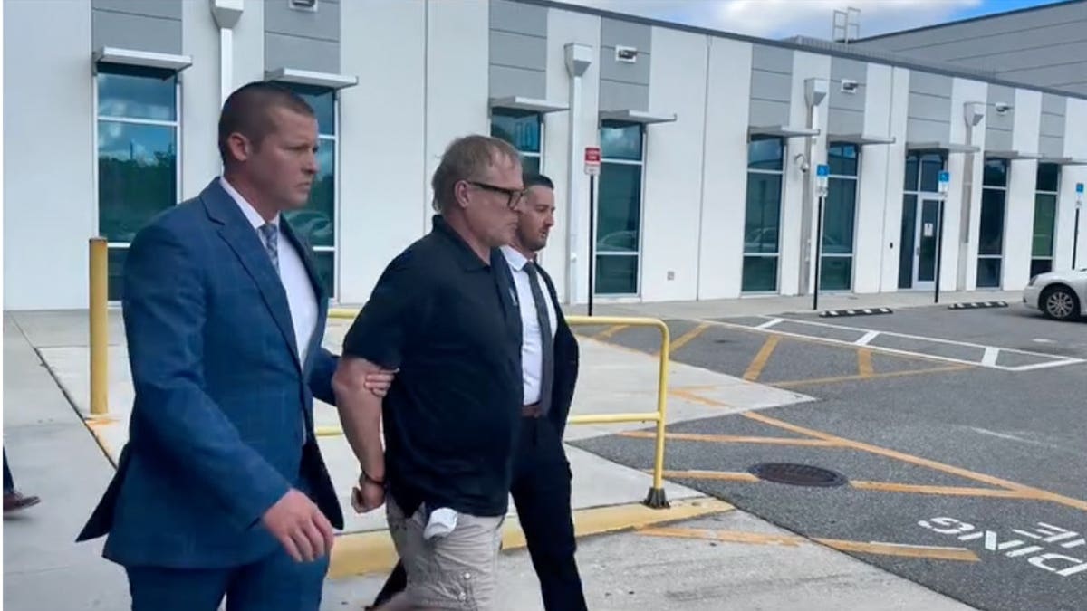 Dale Smith Jr., wearing a black polo shirt and khaki shorts, walks in handcuffs between two plainclothes officers outside a building.