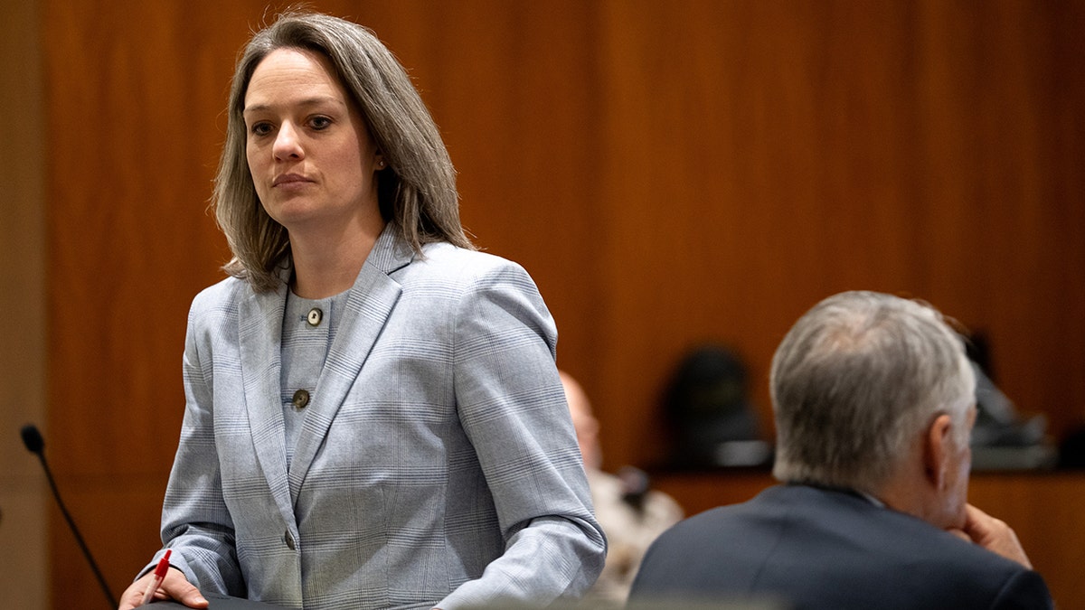 Staci Visser standing in a courtroom wearing a light blue plaid blazer and holding a red pen.