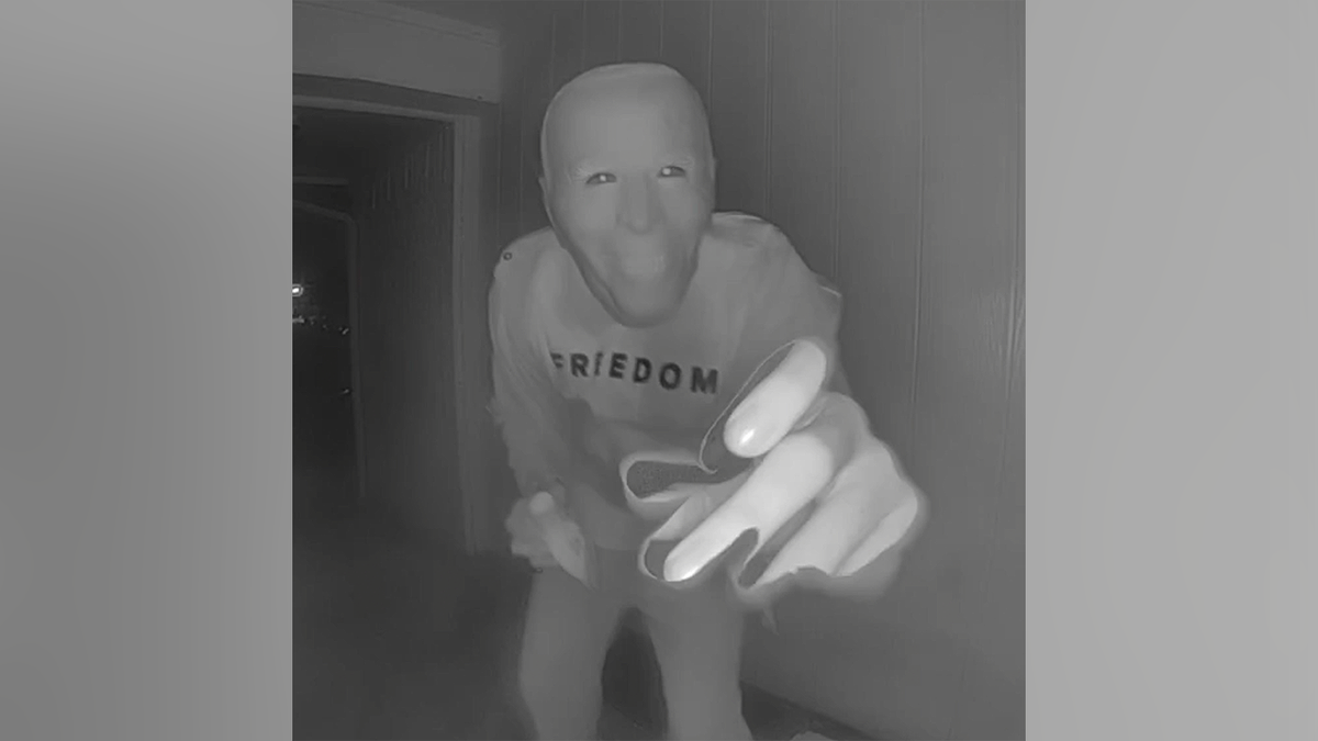 Person wearing a Joe Biden mask reaches toward a doorbell camera while holding a banana