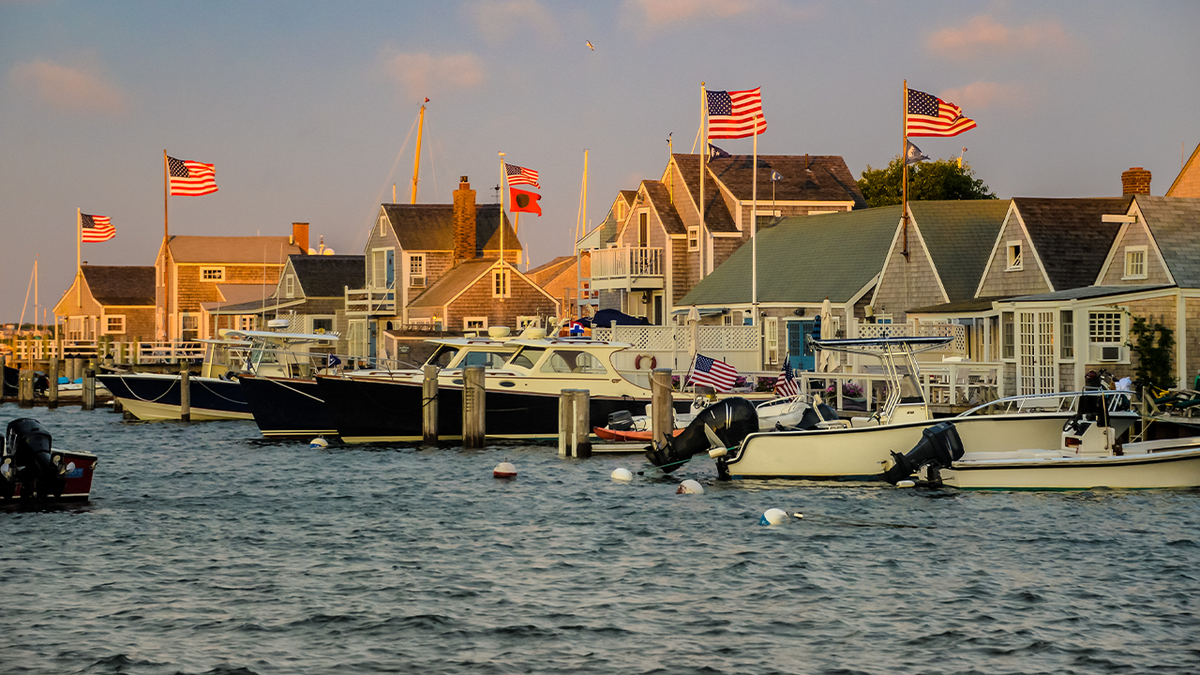 Boats docked in a marina in Nantucket, in front of a row of waterfront houses, many which are flying American flags